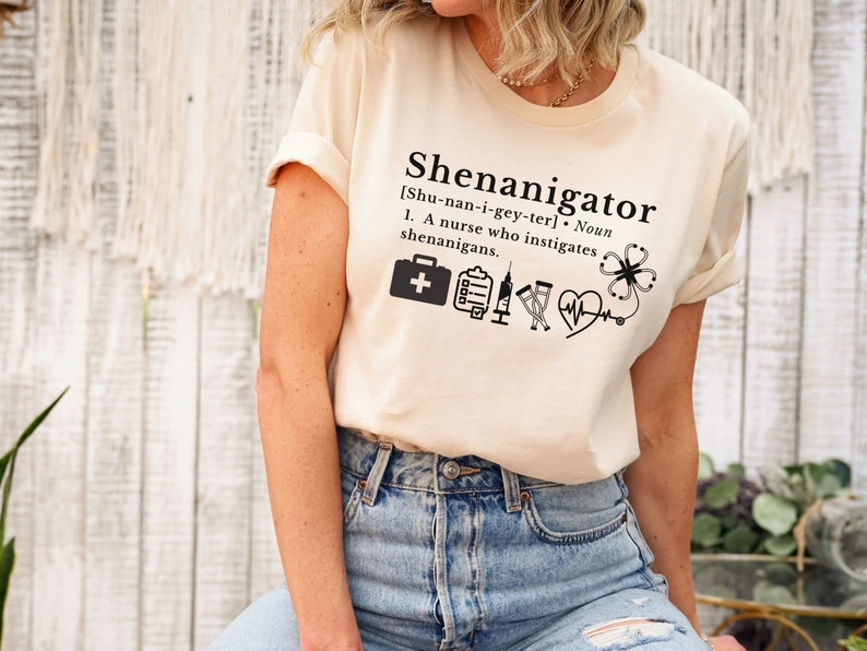 May include: A beige t-shirt with the text "Shenanigator [Shu-nan-i-gey-ter]. Noun 1. A nurse who instigates shenanigans." and a series of medical icons including a first aid kit, a clipboard, a syringe, a vial, a heart rate monitor, and a four-leaf clover.