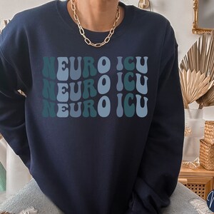 Pode incluir: Camisola azul marinho com o texto "NEURO ICU" repetido tr&ecirc;s vezes em uma fonte retro ondulada. O texto est&aacute; em tons de azul e verde.