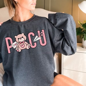 Peut inclure: Un sweat-shirt gris avec un motif graphique rose repr&eacute;sentant le mot "POCU" avec un ours en peluche tenant une lettre et un avion rose.