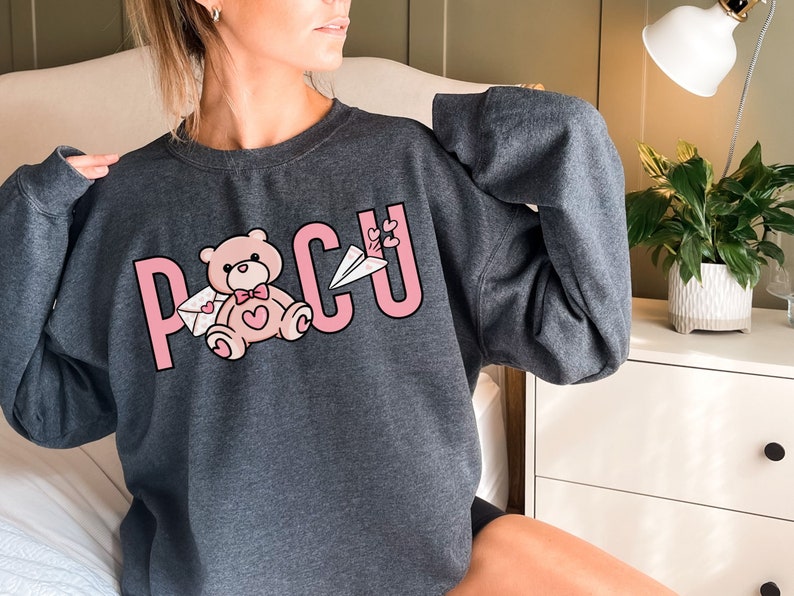 Peut inclure: Un sweat-shirt gris avec un motif graphique rose repr&eacute;sentant le mot "POCU" avec un ours en peluche tenant une lettre et un avion rose.