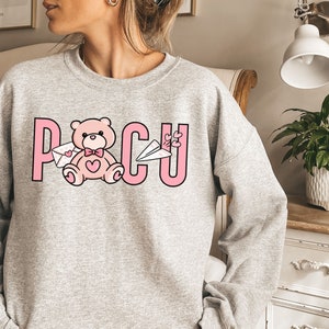 Peut inclure: Un sweat-shirt gris avec un motif rose repr&eacute;sentant un ours en peluche tenant une lettre et le mot "POCU" avec des c&oelig;urs.