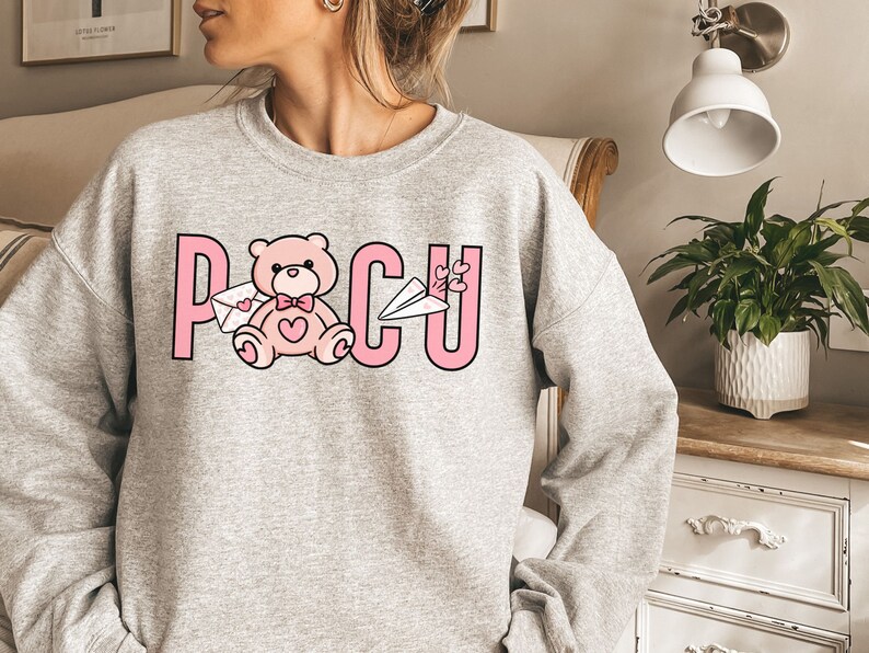 Peut inclure: Un sweat-shirt gris avec un motif rose repr&eacute;sentant un ours en peluche tenant une lettre et le mot "POCU" avec des c&oelig;urs.