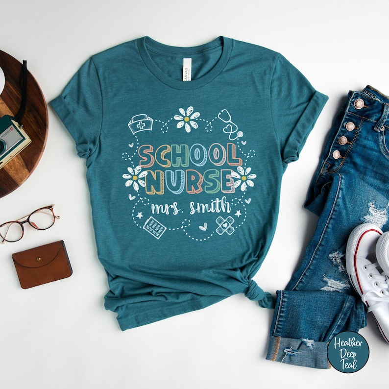 Puede incluir: Camiseta de color verde azulado con las palabras "SCHOOL NURSE mrs. smith" en letras coloridas y un dise&ntilde;o floral. La camiseta se combina con vaqueros azules y zapatillas blancas. La camiseta es de material suave.