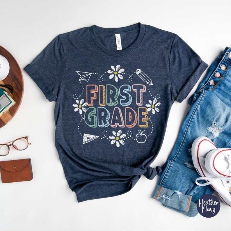 May include: A navy blue t-shirt with the words "FIRST GRADE" in colorful letters, surrounded by daisies, a pencil, a paper airplane, a ruler, and an apple. The shirt is paired with blue jeans and white sneakers.
