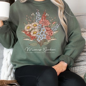 May include: A green sweatshirt with a floral design and the text "Mimi's Garden" printed on the front. The flowers are in shades of pink, white, and blue.
