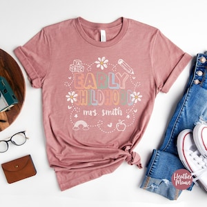 Pode incluir: T-shirt cor de malva com a frase "EARLY CHILDHOOD mrs. smith" em uma fonte colorida e divertida. O design inclui um arco-&iacute;ris, uma ma&ccedil;&atilde;, um l&aacute;pis e margaridas. A camiseta &eacute; combinada com jeans azuis e t&ecirc;nis brancos.