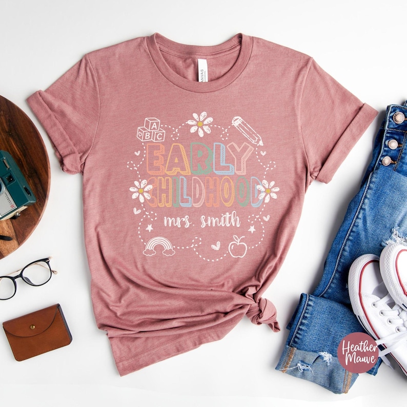 Pode incluir: T-shirt cor de malva com a frase "EARLY CHILDHOOD mrs. smith" em uma fonte colorida e divertida. O design inclui um arco-&iacute;ris, uma ma&ccedil;&atilde;, um l&aacute;pis e margaridas. A camiseta &eacute; combinada com jeans azuis e t&ecirc;nis brancos.