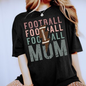 Op de afbeelding: Zwart T-shirt met een distressed grafisch ontwerp met de tekst "Football Mom" met een bliksem en een voetbalafbeelding.