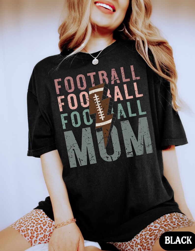 Op de afbeelding: Zwart T-shirt met een distressed grafisch ontwerp met de tekst "Football Mom" met een bliksem en een voetbalafbeelding.