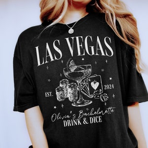 May include: Black t-shirt with white text that says "Las Vegas" and "Olivia's Bachelorette Drink & Dice" with a graphic of a cocktail glass, playing cards, dice, and chips. The year 2024 and "Est." are also printed on the shirt.