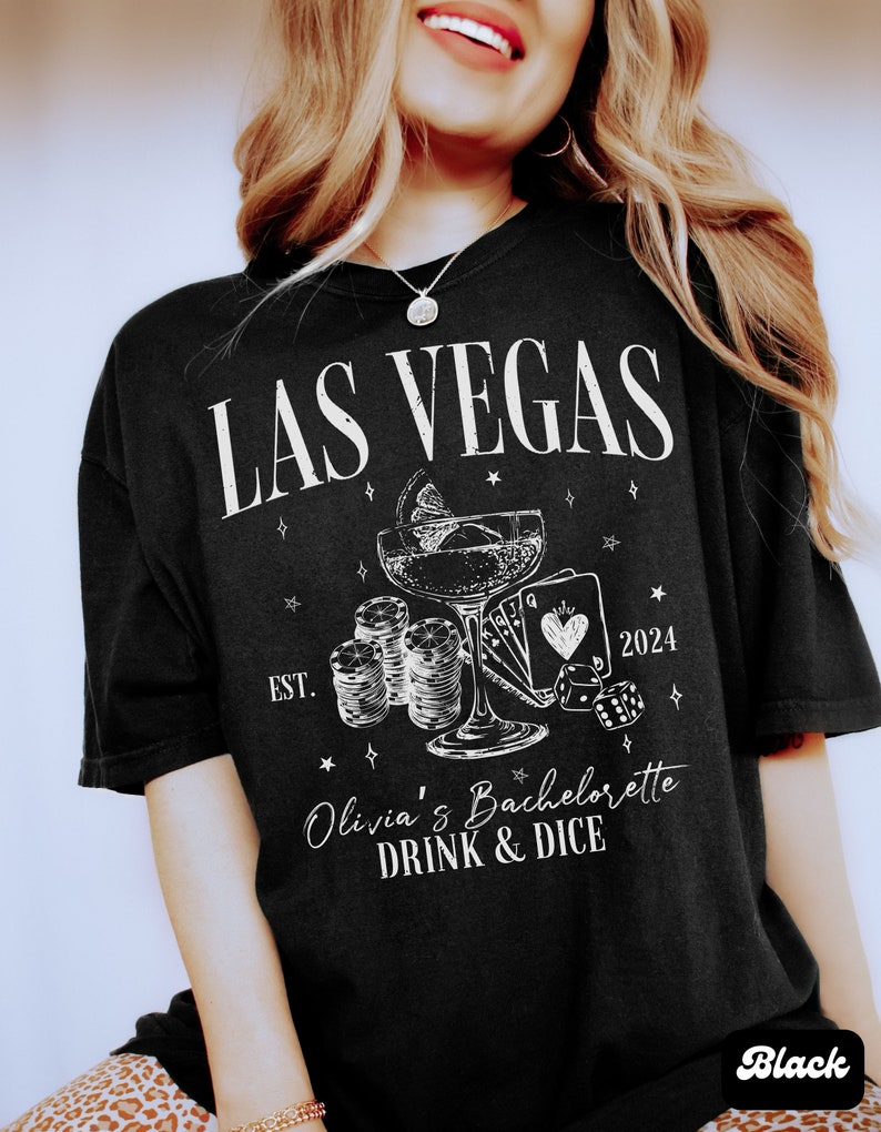 May include: Black t-shirt with white text that says "Las Vegas" and "Olivia's Bachelorette Drink & Dice" with a graphic of a cocktail glass, playing cards, dice, and chips. The year 2024 and "Est." are also printed on the shirt.