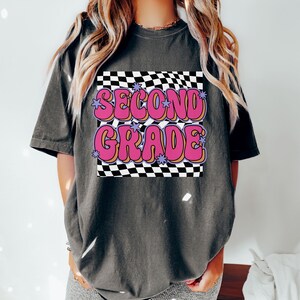 以下が含まれることがあります： 黒っぽいグレーのTシャツに、市松模様と「SECOND GRADE」という文字がピンクと紫の文字で書かれている。