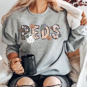 May include: A heather gray sweatshirt with the word "PEDS" in a Halloween-themed design. The letters are decorated with pumpkins, ghosts, and floral patterns. A black mug is held in the hand.