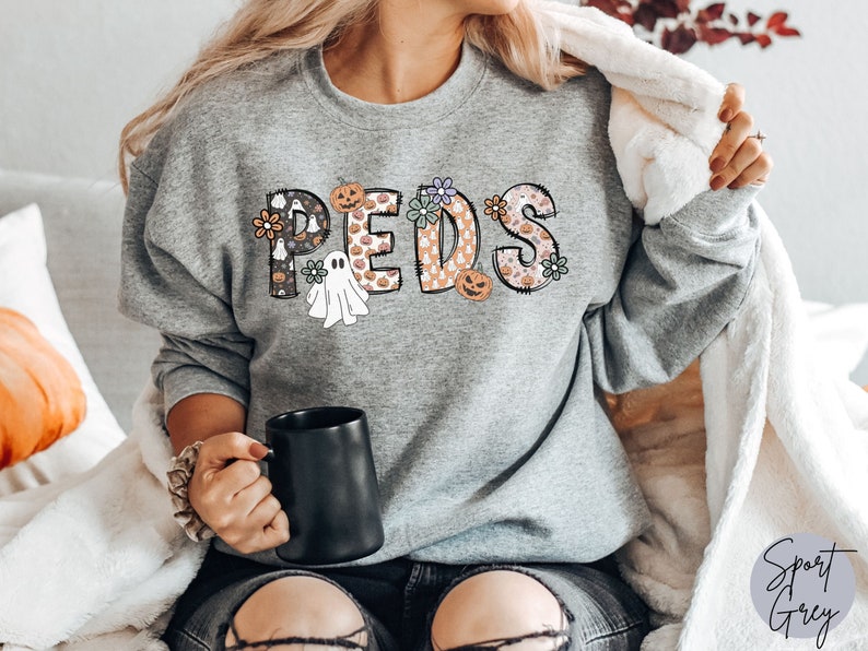 May include: A heather gray sweatshirt with the word "PEDS" in a Halloween-themed design. The letters are decorated with pumpkins, ghosts, and floral patterns. A black mug is held in the hand.
