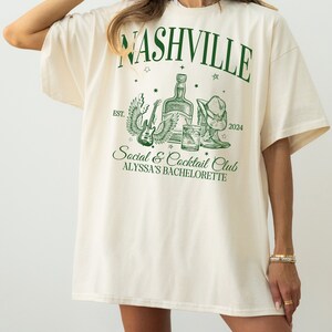 K&ouml;nnte beinhalten: Ein cremefarbenes T-Shirt mit einem gr&uuml;nen Grafikdesign, auf dem "Nashville Social & Cocktail Club Alyssa's Bachelorette" steht, mit einer Flasche Alkohol, einer Gitarre und einem Cowboyhut.