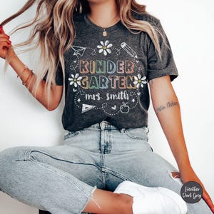 May include: A dark heather grey t-shirt with the text "Kindergarten" in rainbow colors and the text "mrs. smith" in white. The shirt has a variety of hand-drawn school-themed graphics, including a pencil, a paper airplane, a daisy, a ruler, an apple, and hearts.