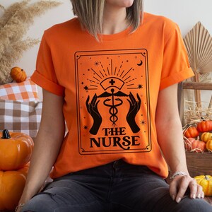 May include: Orange t-shirt featuring a black graphic of a tarot card that reads "The Nurse" with a medical caduceus symbol.