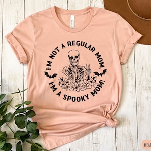 May include: A peach-colored t-shirt with a black graphic of a skeleton with a peace sign and the text "I'm not a regular mom. I'm a spooky mom!"