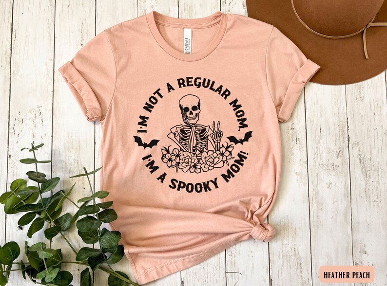 May include: A peach-colored t-shirt with a black graphic of a skeleton with a peace sign and the text "I'm not a regular mom. I'm a spooky mom!"