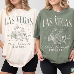 May include: Two white t-shirts with a green graphic design. The graphic features a cocktail glass, dice, and playing cards. The text on the shirts reads "Las Vegas Est. 2024 Olivia's Bachelorette Drink & Dice". The shirts are on models wearing black shorts. The shirt on the left is ivory and the shirt on the right is moss.