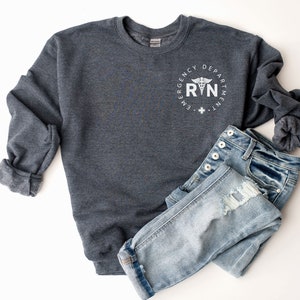 Peut inclure: Sweat-shirt gris fonc&eacute; chin&eacute; avec un graphique circulaire blanc qui dit "Emergency Department RN" avec un symbole de caduc&eacute;e.