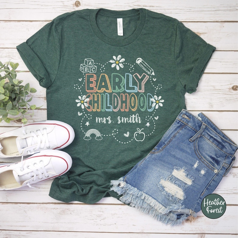 Pode incluir: Uma camiseta verde com as palavras "EARLY CHILDHOOD mrs. smith" em letras coloridas e gr&aacute;ficos. A camiseta &eacute; combinada com shorts jeans e t&ecirc;nis brancos. A camiseta tem mangas enroladas e &eacute; exibida em uma superf&iacute;cie de madeira clara.