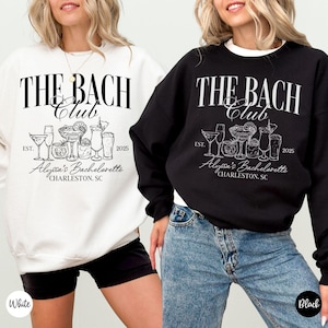 K&ouml;nnte beinhalten: Zwei wei&szlig;e und schwarze Sweatshirts mit dem Text "The Bach Club" und einer Grafik von Cocktailgl&auml;sern. Der Text "EST. 2025" und "Alyssa's Bachelorette" sind ebenfalls auf den Sweatshirts enthalten. Das wei&szlig;e Sweatshirt wird von einer Person mit schwarzen Shorts getragen und das schwarze Sweatshirt von einer Person mit blauen Jeans.
