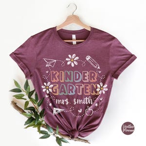 May include: A maroon t-shirt with the text "KINDERGARTEN" in rainbow colors and "mrs. smith" in white. The shirt has a design of flowers, a pencil, a paper airplane, a ruler, an apple, and stars.