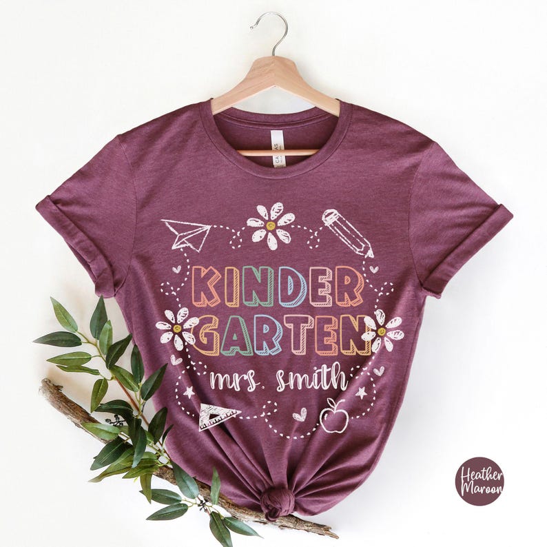 May include: A maroon t-shirt with the text "KINDERGARTEN" in rainbow colors and "mrs. smith" in white. The shirt has a design of flowers, a pencil, a paper airplane, a ruler, an apple, and stars.