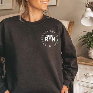 Peut inclure: Sweat-shirt noir avec un graphique blanc d'un symbole m&eacute;dical et le texte "Emergency Department RN".