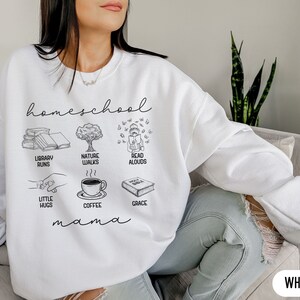 Puede incluir: Sudadera blanca con el texto "homeschool mama" e ilustraciones de libros, un &aacute;rbol, un ni&ntilde;o leyendo, caf&eacute; y un abrazo.