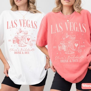 May include: Two white t-shirts with a pink design. The design features the text "LAS VEGAS" with a cocktail glass, dice, and playing cards. The text "Olivia's Bachelorette DRINK & DICE" is below the design. One shirt is ivory and the other is watermelon.