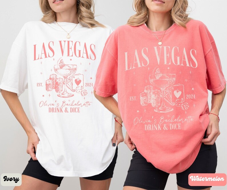 May include: Two white t-shirts with a pink design. The design features the text "LAS VEGAS" with a cocktail glass, dice, and playing cards. The text "Olivia's Bachelorette DRINK & DICE" is below the design. One shirt is ivory and the other is watermelon.