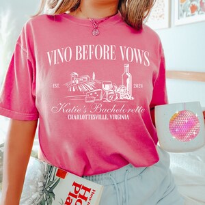 May include: Pink t-shirt with the text "Vino Before Vows" and a graphic of a vineyard, wine bottle, grapes, and cheese. The text "Est. 2024" and "Katie's Bachelorette Charlottesville, Virginia" are also on the shirt.