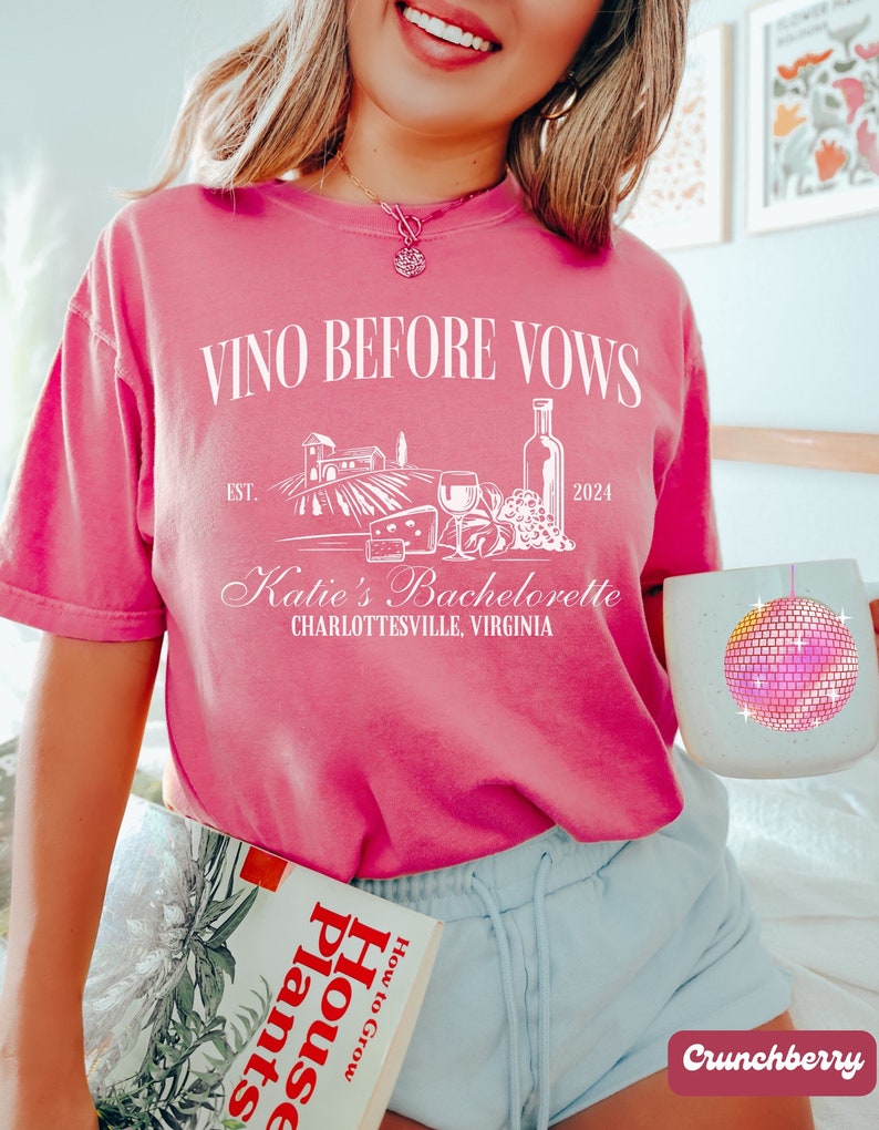 May include: Pink t-shirt with the text "Vino Before Vows" and a graphic of a vineyard, wine bottle, grapes, and cheese. The text "Est. 2024" and "Katie's Bachelorette Charlottesville, Virginia" are also on the shirt.