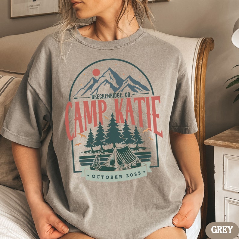 May include: A grey t-shirt with a vintage-style graphic of a mountain range, a tent, and trees. The text "Camp Katie" is printed in red and blue, with the words "Breckenridge, Co." and "October 2023" printed below.