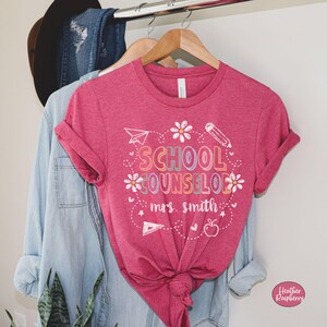 May include: A pink t-shirt with rolled-up sleeves, featuring the words "SCHOOL COUNSELOR" in colorful, outlined letters. The design includes a paper airplane, pencil, flowers, and an apple. The name "mrs. smith" is printed below the main text.