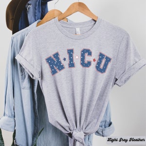 May include: A light grey heather t-shirt with the text "NICU" in blue and red lettering with white stars. The letters are outlined in red.