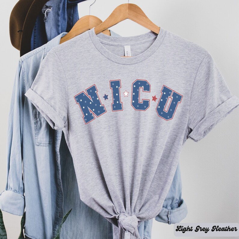 May include: A light grey heather t-shirt with the text "NICU" in blue and red lettering with white stars. The letters are outlined in red.