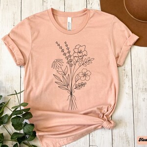 K&ouml;nnte beinhalten: Ein pfirsichfarbenes T-Shirt mit einer schwarzen Linienzeichnung eines Blumenstrau&szlig;es. Das Shirt ist aus einem weichen, bequemen Material gefertigt und hat eine lockere Passform.
