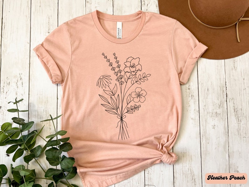 K&ouml;nnte beinhalten: Ein pfirsichfarbenes T-Shirt mit einer schwarzen Linienzeichnung eines Blumenstrau&szlig;es. Das Shirt ist aus einem weichen, bequemen Material gefertigt und hat eine lockere Passform.