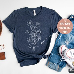 K&ouml;nnte beinhalten: Ein heidefarbenes marineblaues T-Shirt mit einem wei&szlig;en Blumenmuster und dem Wort "Grandma" auf der Vorderseite. Das Shirt ist auf einem wei&szlig;en Hintergrund mit einer Jeans und wei&szlig;en Turnschuhen abgebildet.