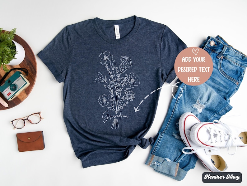 K&ouml;nnte beinhalten: Ein heidefarbenes marineblaues T-Shirt mit einem wei&szlig;en Blumenmuster und dem Wort "Grandma" auf der Vorderseite. Das Shirt ist auf einem wei&szlig;en Hintergrund mit einer Jeans und wei&szlig;en Turnschuhen abgebildet.