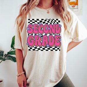 以下が含まれることがあります： 白地にチェック柄のTシャツに、ピンクと紫の文字で「SECOND GRADE」と星が書かれている。黒いショートパンツを履いた人が着ている。