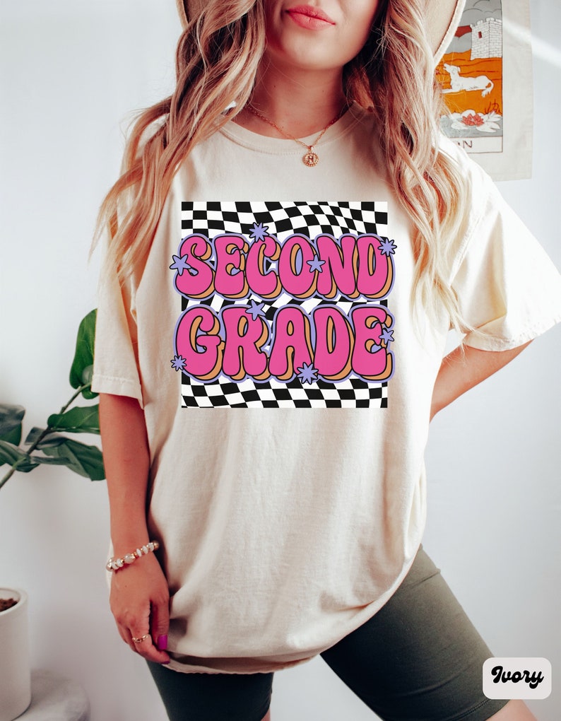 以下が含まれることがあります： 白地にチェック柄のTシャツに、ピンクと紫の文字で「SECOND GRADE」と星が書かれている。黒いショートパンツを履いた人が着ている。
