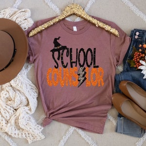 May include: A mauve t-shirt with a black witch hat and the text "School Counselor" in orange glitter.