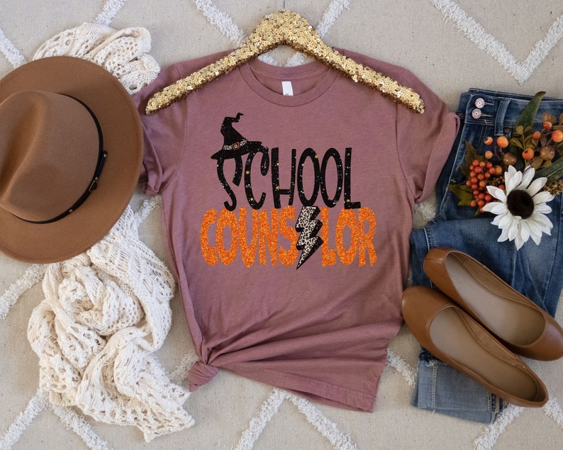 May include: A mauve t-shirt with a black witch hat and the text "School Counselor" in orange glitter.