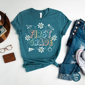 May include: Teal-colored t-shirt with the words "FIRST GRADE" in colorful, outlined letters. The design includes white daisies, a paper airplane, a pencil, a ruler, and an apple. The shirt is paired with blue jeans and white sneakers.