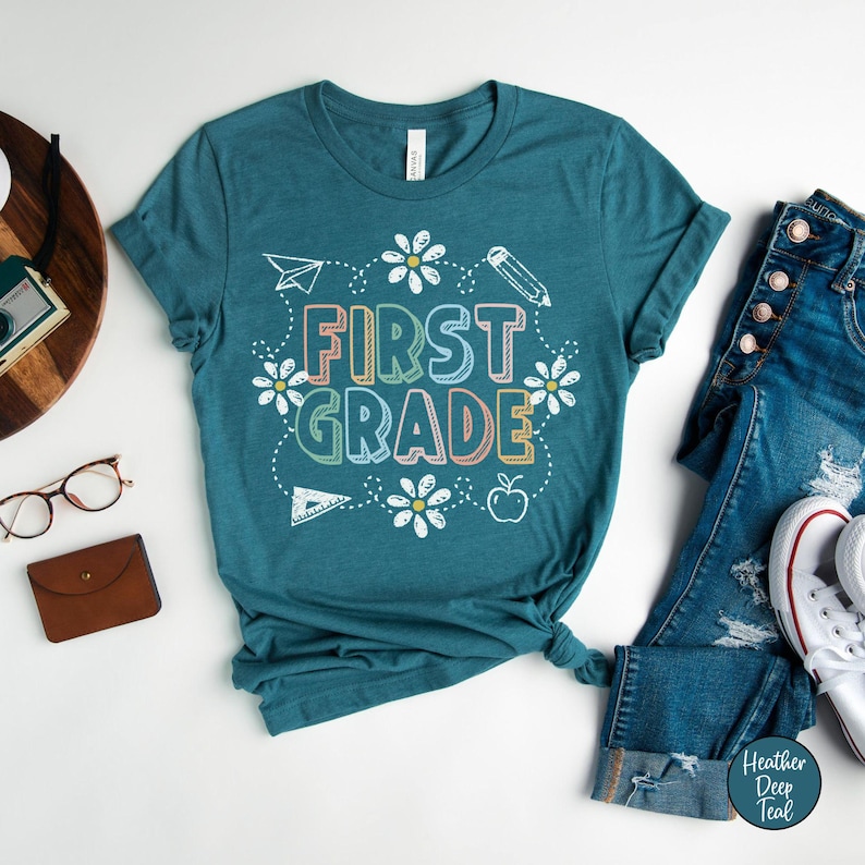 May include: Teal-colored t-shirt with the words "FIRST GRADE" in colorful, outlined letters. The design includes white daisies, a paper airplane, a pencil, a ruler, and an apple. The shirt is paired with blue jeans and white sneakers.