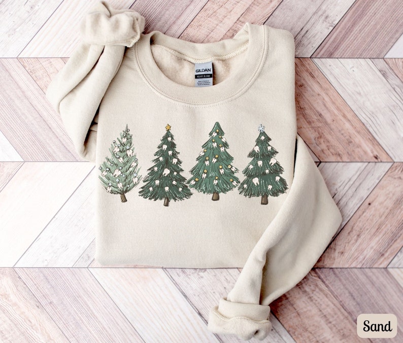K&ouml;nnte beinhalten: Ein beiger Pullover mit einem Aufdruck von vier Weihnachtsb&auml;umen, die mit Z&auml;hnen verziert sind. Die B&auml;ume sind gr&uuml;n mit wei&szlig;en Z&auml;hnen und gelben Ornamenten. Der Pullover hat ein Gildan-Etikett.